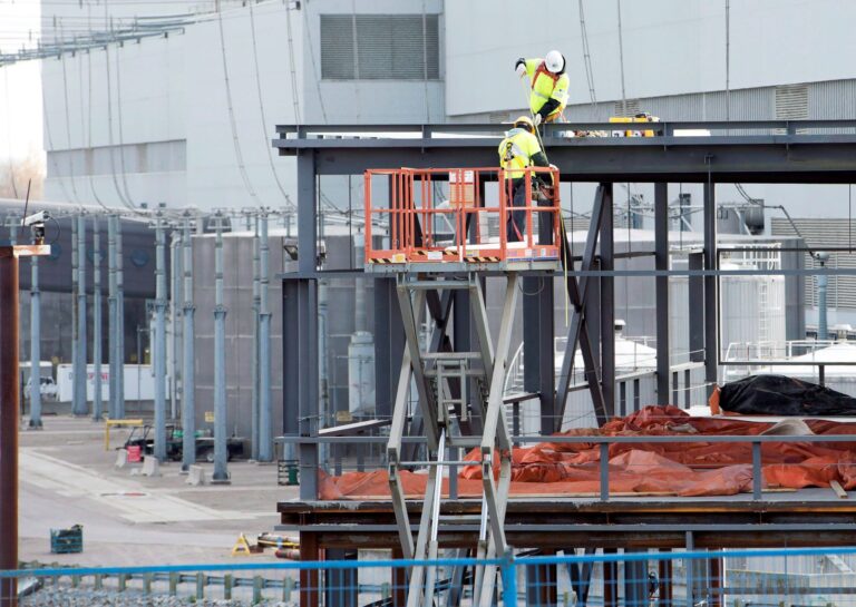 Two workers in hard hats labour on the framing of a building.
