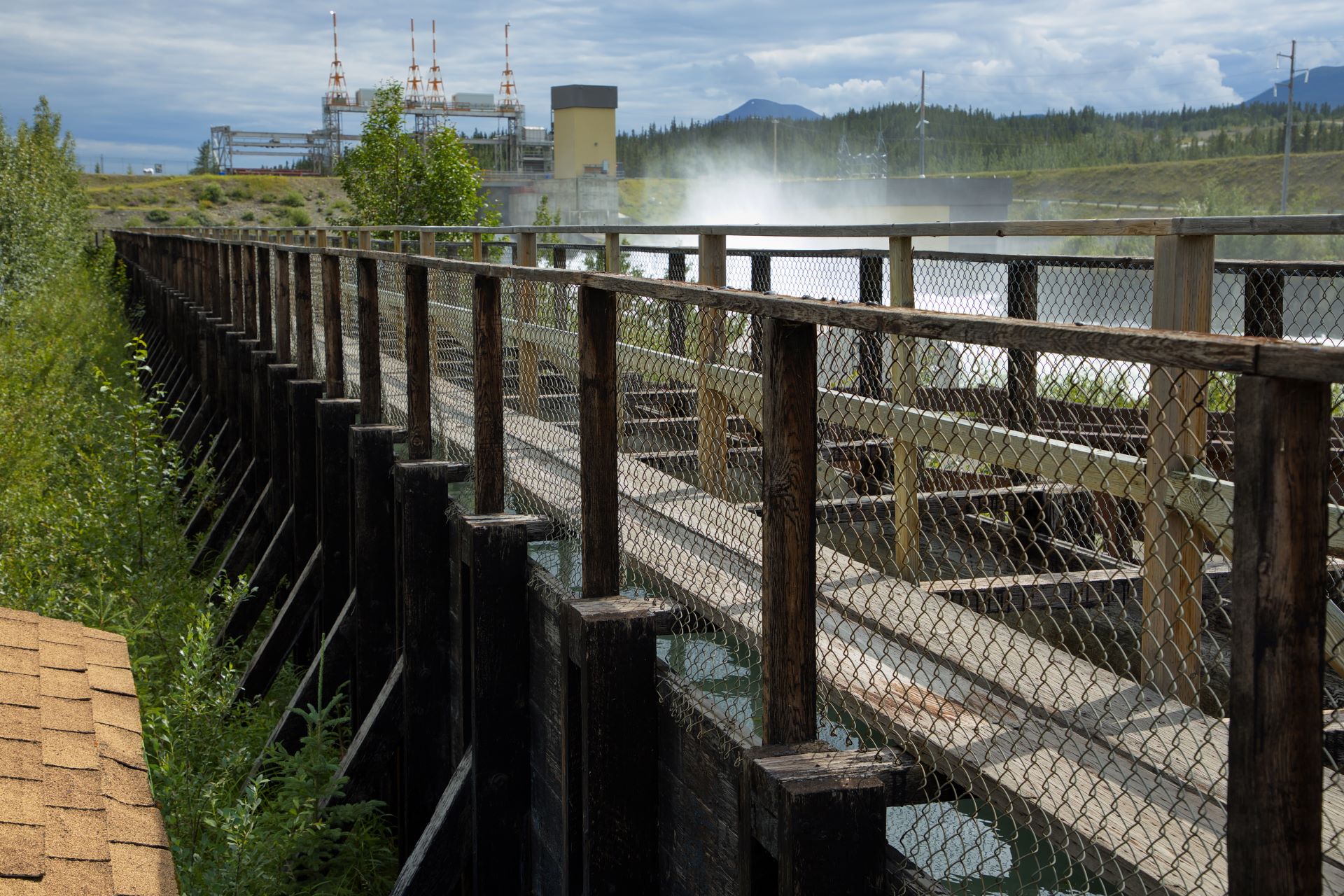 A long wood structure leads to the hydro station in the distance. Water sprays up from below.