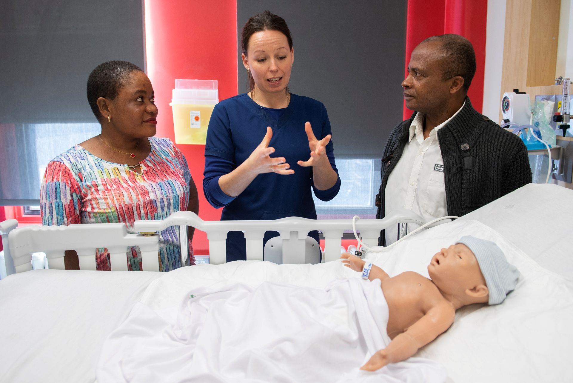 Three people standing over a hospital bed with a dummy baby.
