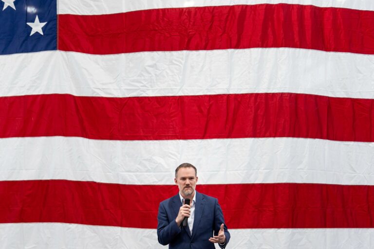 Jamieson Greer stands in front of a huge American flag at a podium.