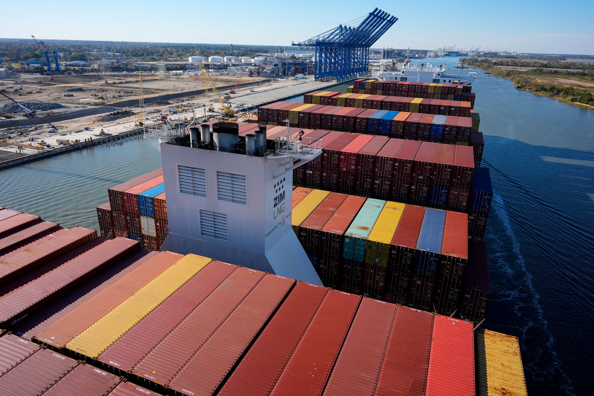 Multi-coloured shipping containers on a narrow river with crane installations along the shore.