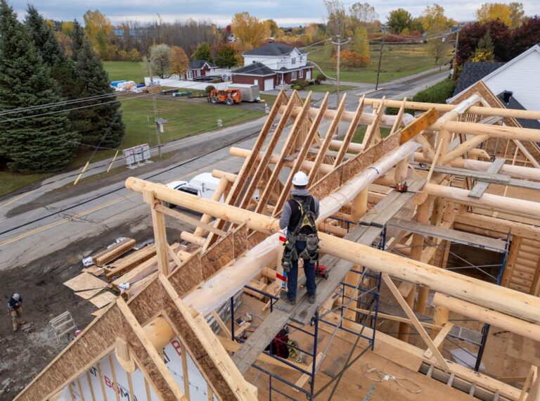 A worker in a white helmet stands on scaffolding near the support beams for a roof.