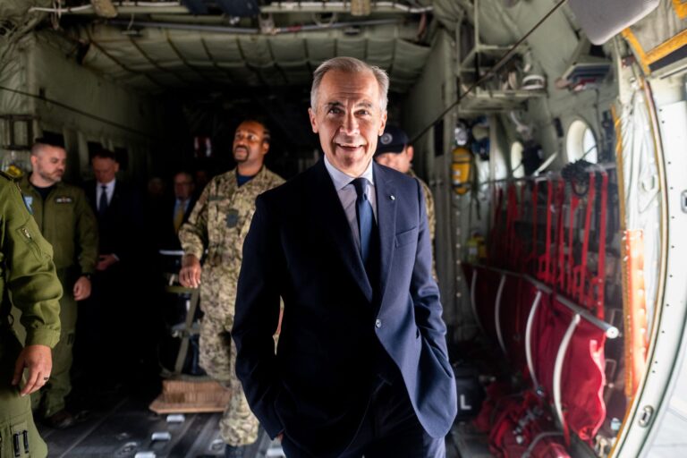 Carney stands smiling, hands in suit-pants pockets, in the cavernous space of a military transport aircraft. Around him are several men in khaki and camouflage uniforms.