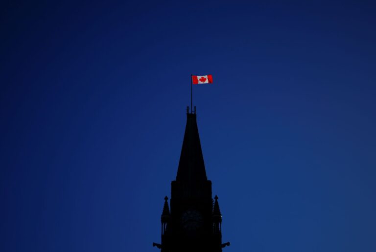 Against a deep blue sky, the parliament Hill building is in silhouette with the sun hitting only the Canadian flag flying from the top of the peace tower.