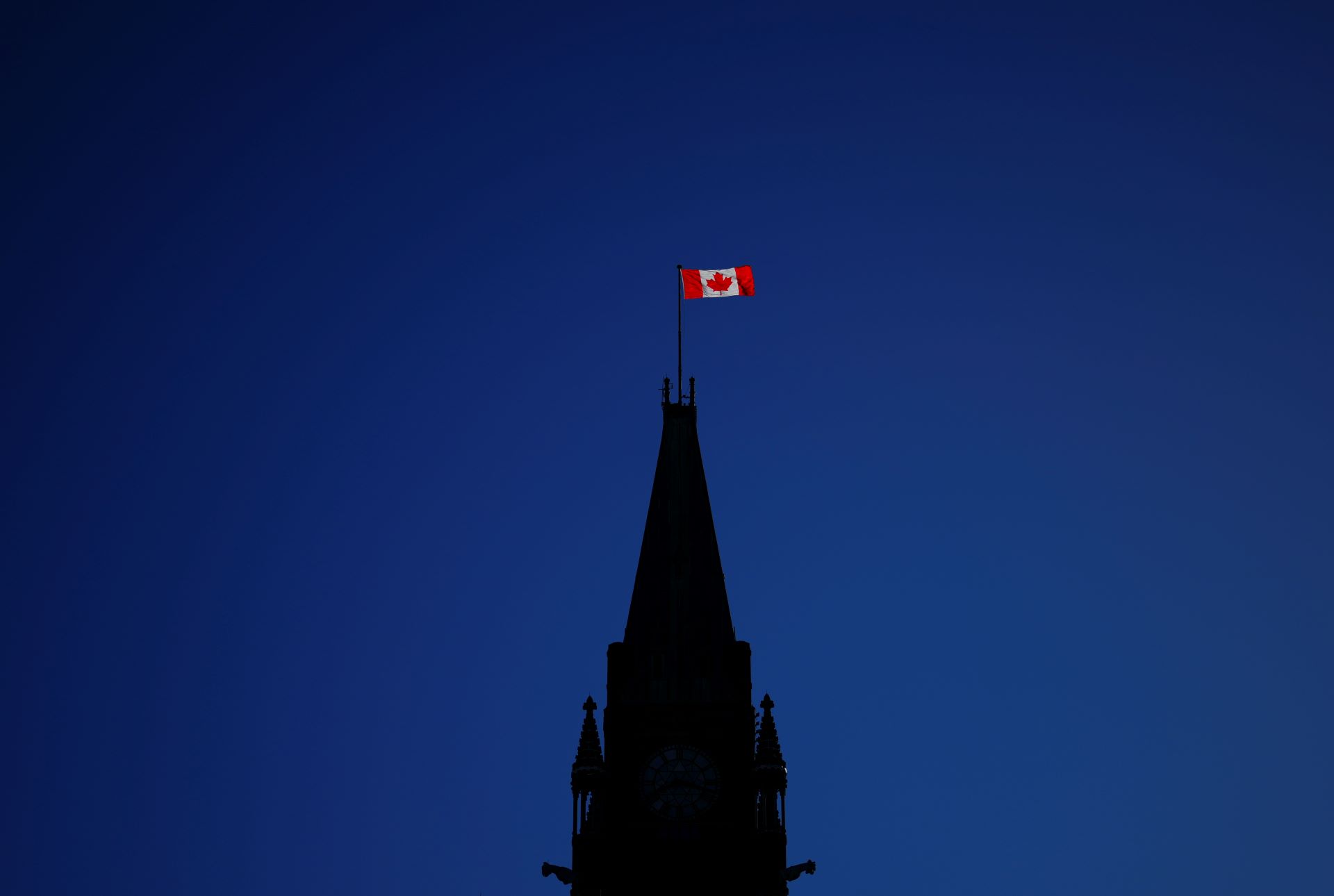 Against a deep blue sky, the parliament Hill building is in silhouette with the sun hitting only the Canadian flag flying from the top of the peace tower.