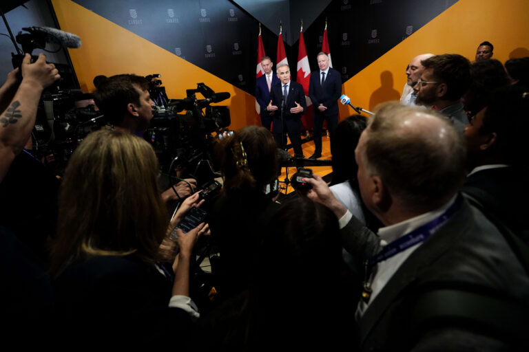 A gaggle of reporters in the foreground with the three men at a podium in the background.