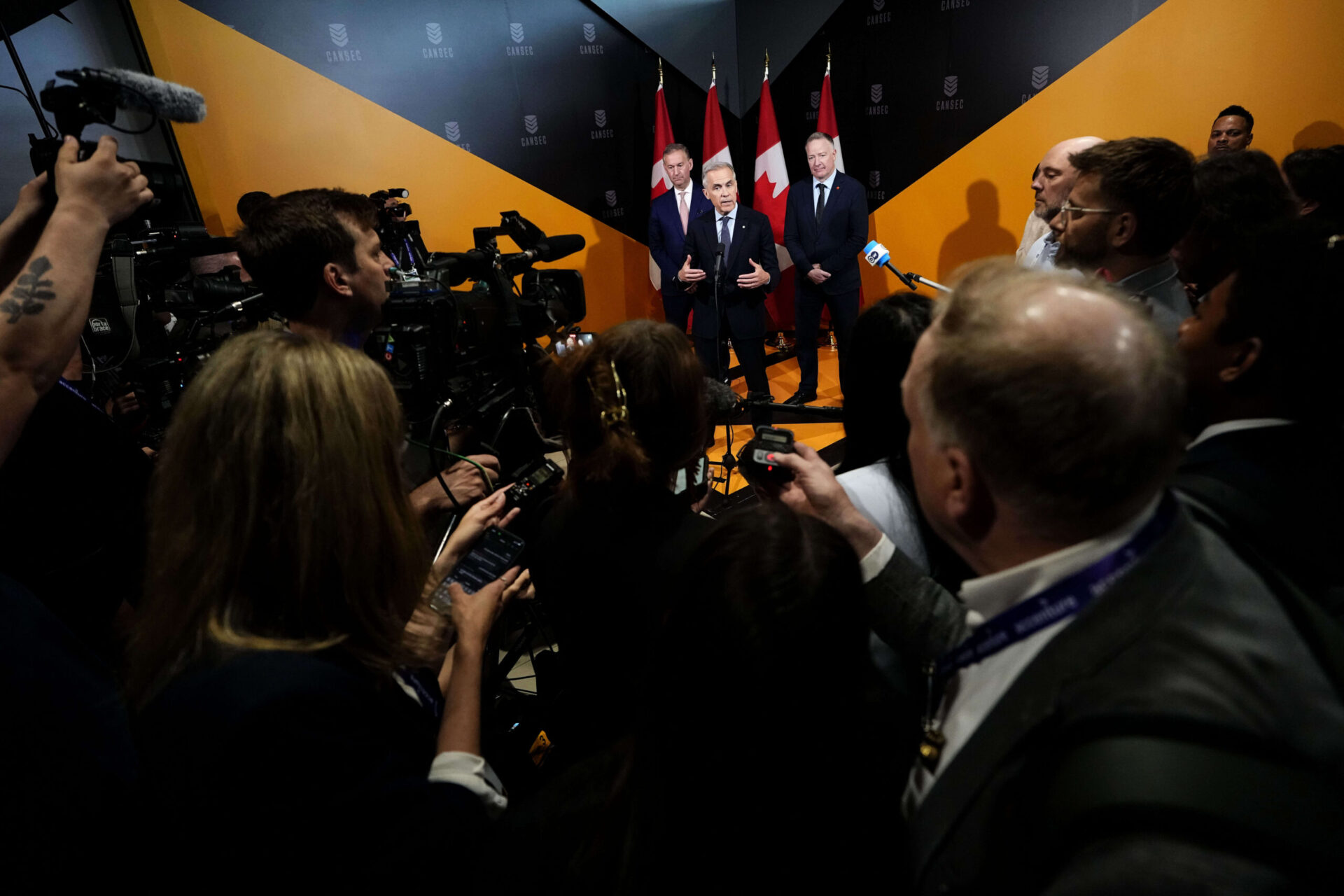 A gaggle of reporters in the foreground with the three men at a podium in the background.