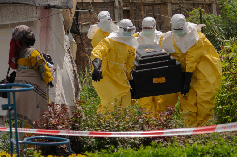Four workers dressed in yellow protective suits with goggles and white head coverings carry a coffin through a patch of short vegetation while a distressed woman looks on. They are all behind red caution tape.