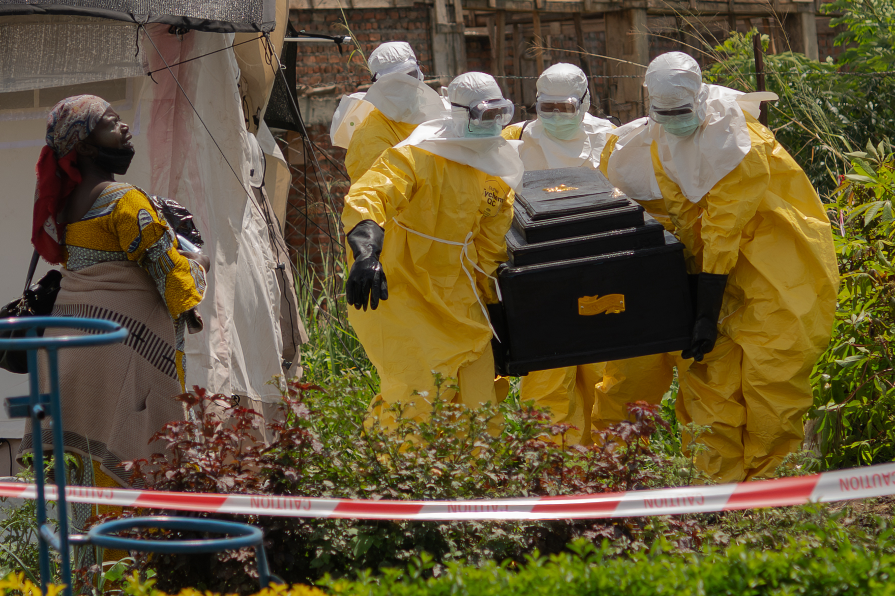 Four workers dressed in yellow protective suits with goggles and white head coverings carry a coffin through a patch of short vegetation while a distressed woman looks on. They are all behind red caution tape.