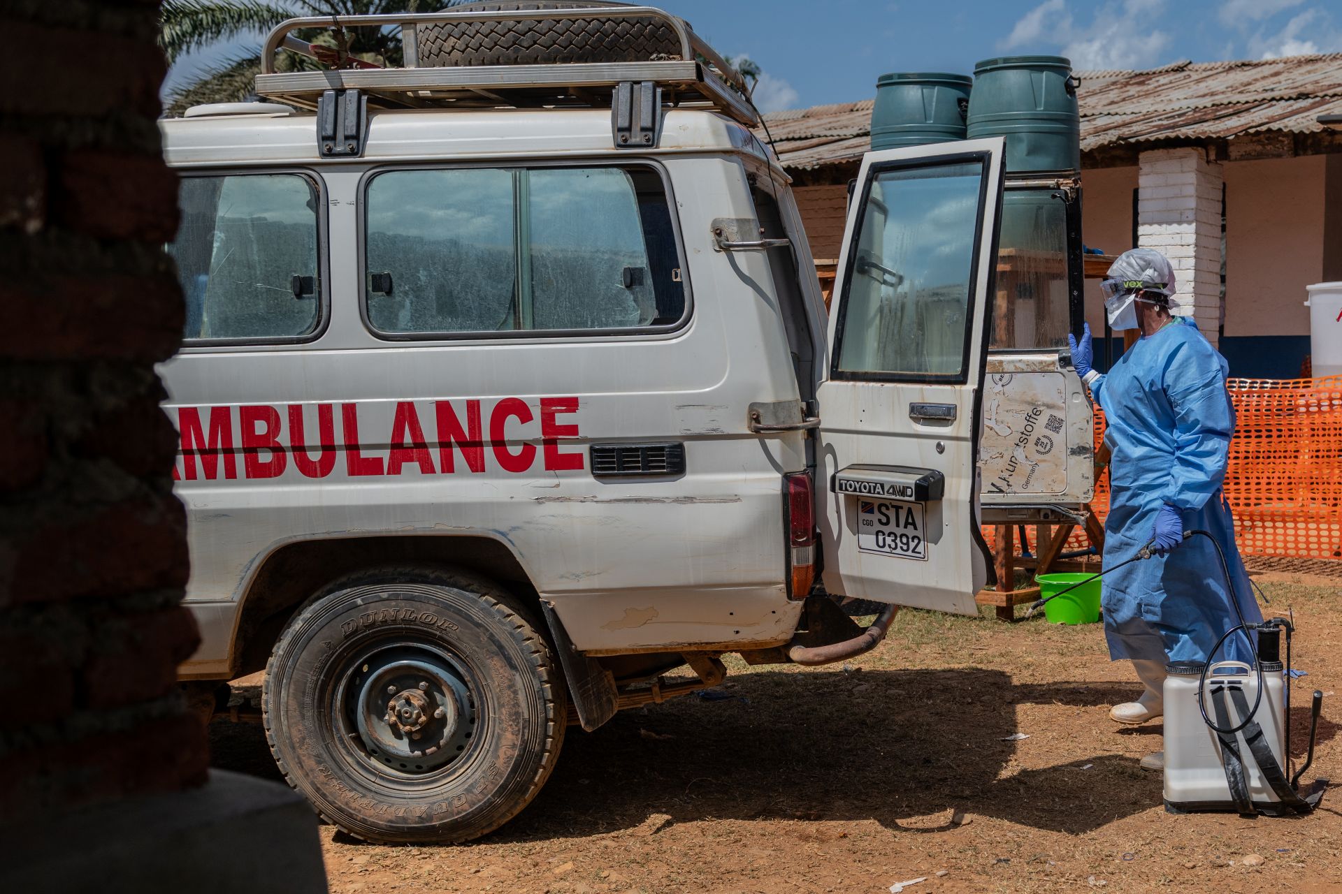 A worker dressed in a blue gown and goggles sprays an grey ambulance Toyota SUV with disinfectant.