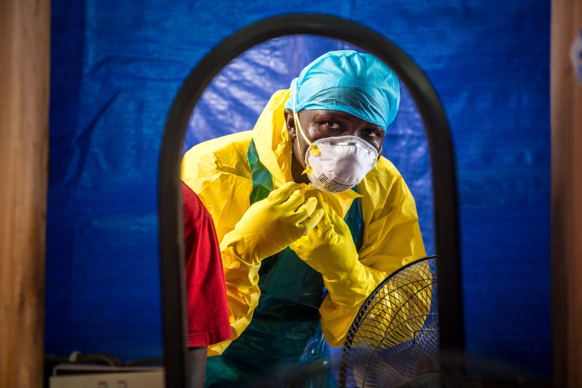 A man in a yellow suit with a white mask and blue head covering peers through a portal into a treatment centre.