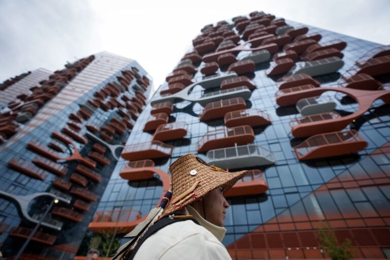 Un homme avec un chapeau, debout devant un immeuble neuf doté de petits balcons.