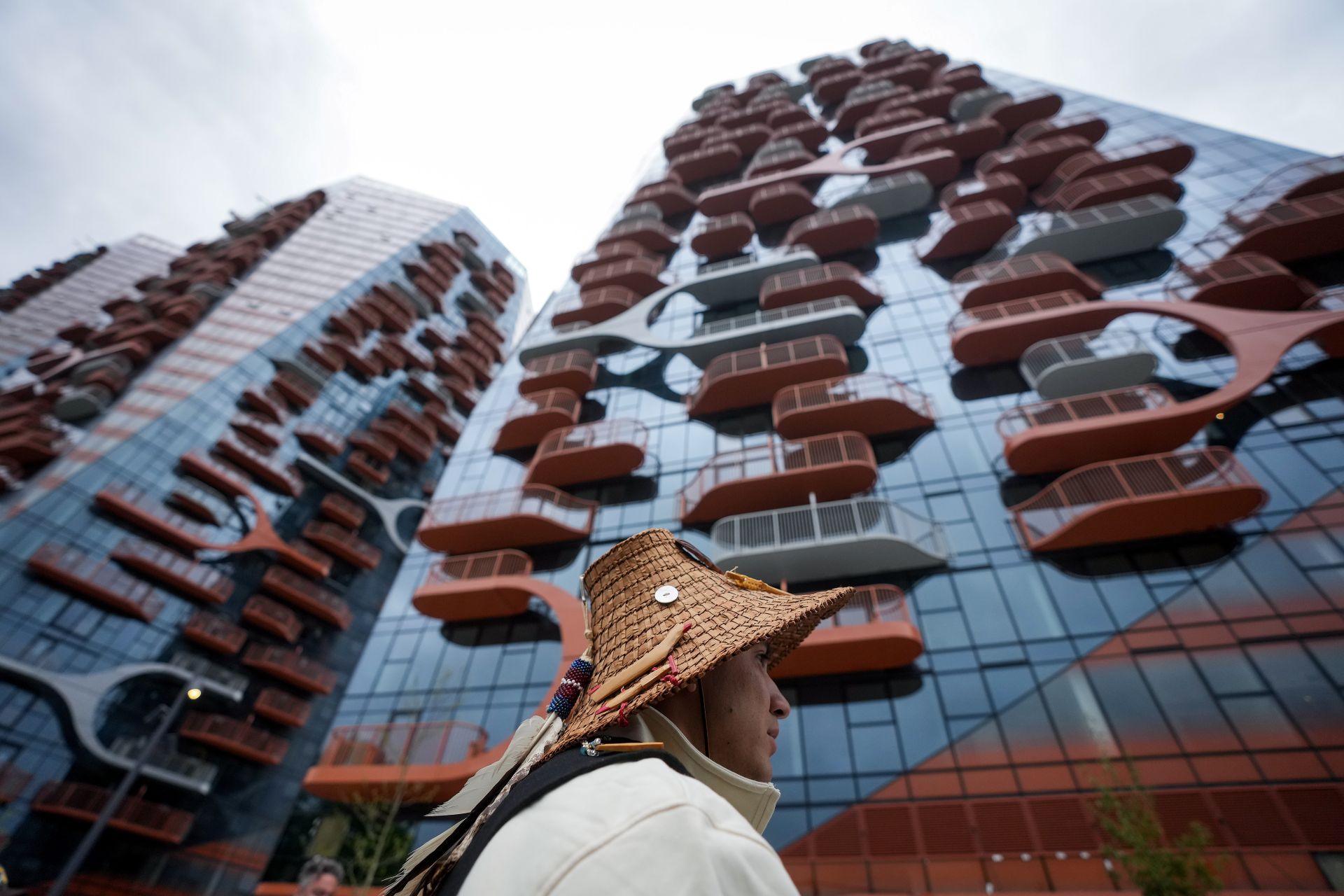 Un homme avec un chapeau, debout devant un immeuble neuf doté de petits balcons.