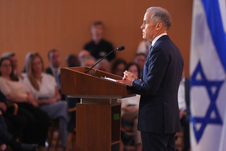 Carney stands at a podium in front of a blue-and-white Jewish flag.