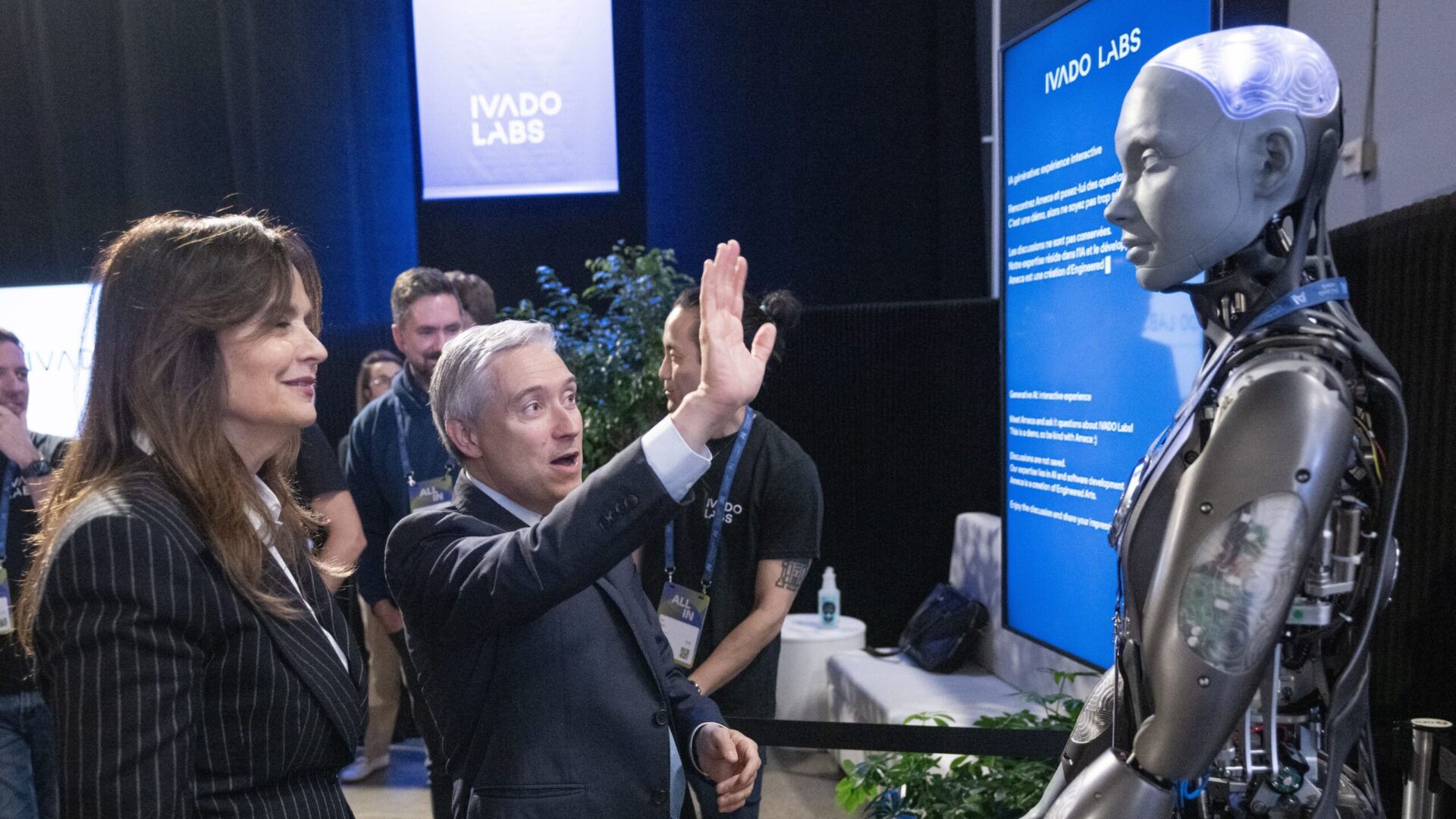 Champagne raises a hand to the robot, which is made of metal but has a human form, while Desmarais looks on.