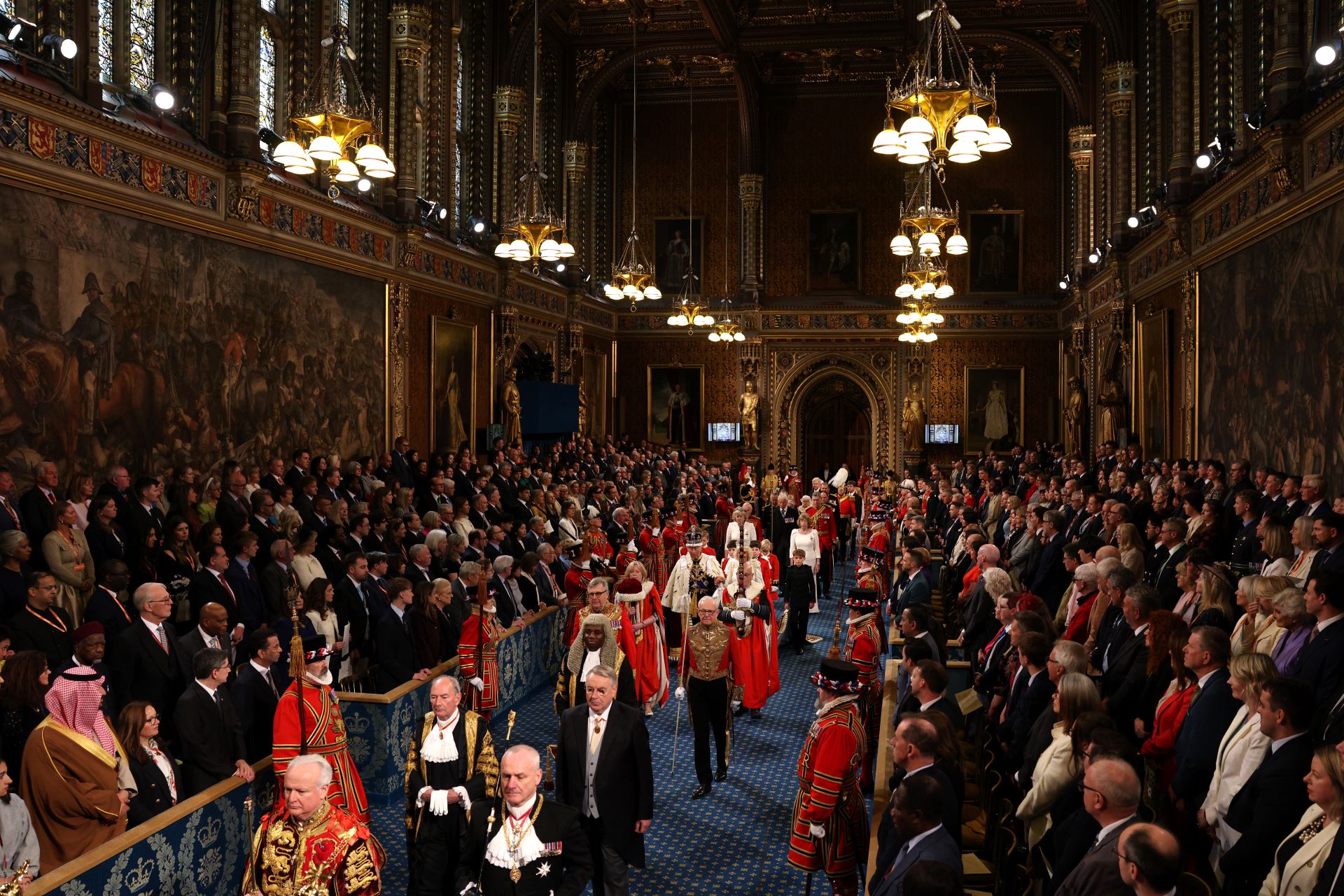 The long room is packed with people on both sides of a central aisle, everyone watching the procession. 