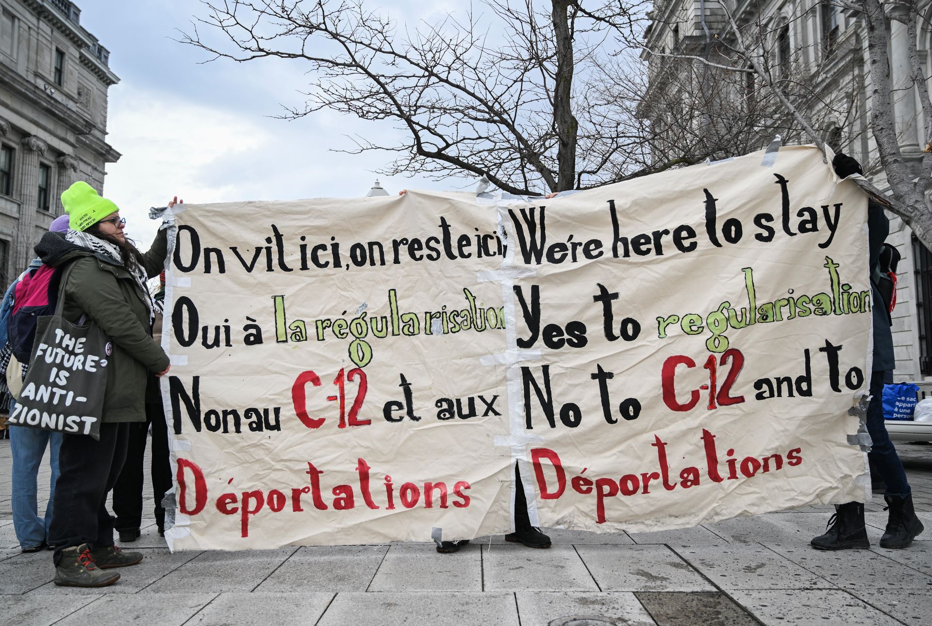 Protesters hold a bilingual banner opposing Bill C-12 and deportations while supporting migrant regularization.