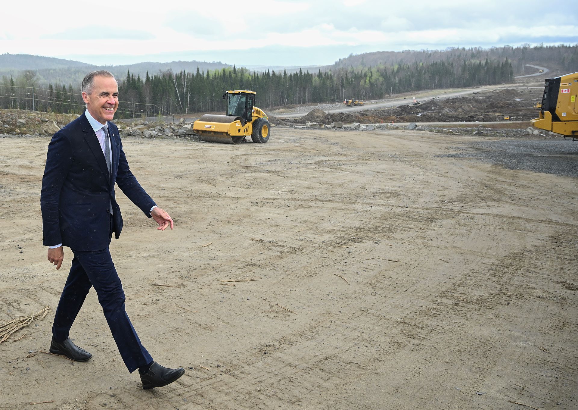 Carney walks across a clearing in the forest. An excavator and roller sit nearby.