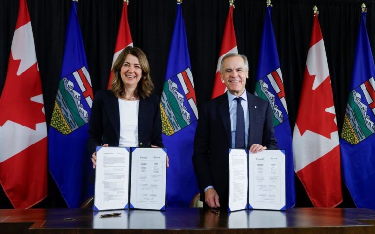 : Danielle Smith (left) and Mark Carney stand posing with copies of their memorandum of understanding with Alberta and Canadian flags in the background.