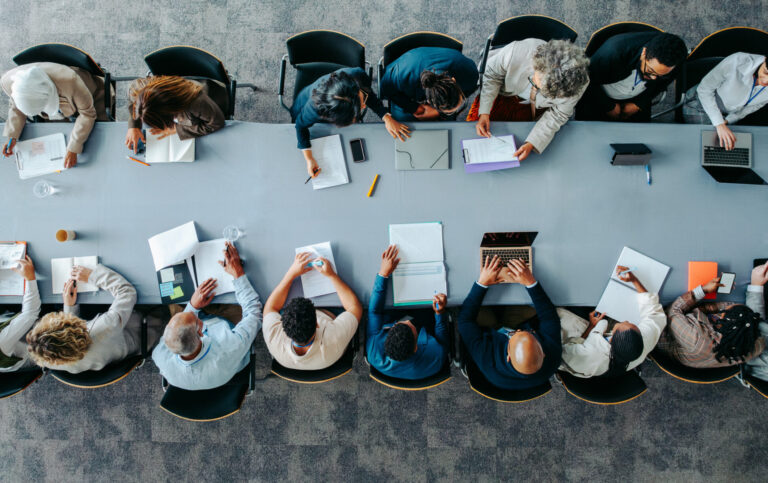 Vue aérienne d’une salle de réunion avec plusieurs personnes assises de part et d’autre d’une longue table, en train de travailler avec des documents, des carnets et des ordinateurs portables, dans un contexte de collaboration professionnelle.