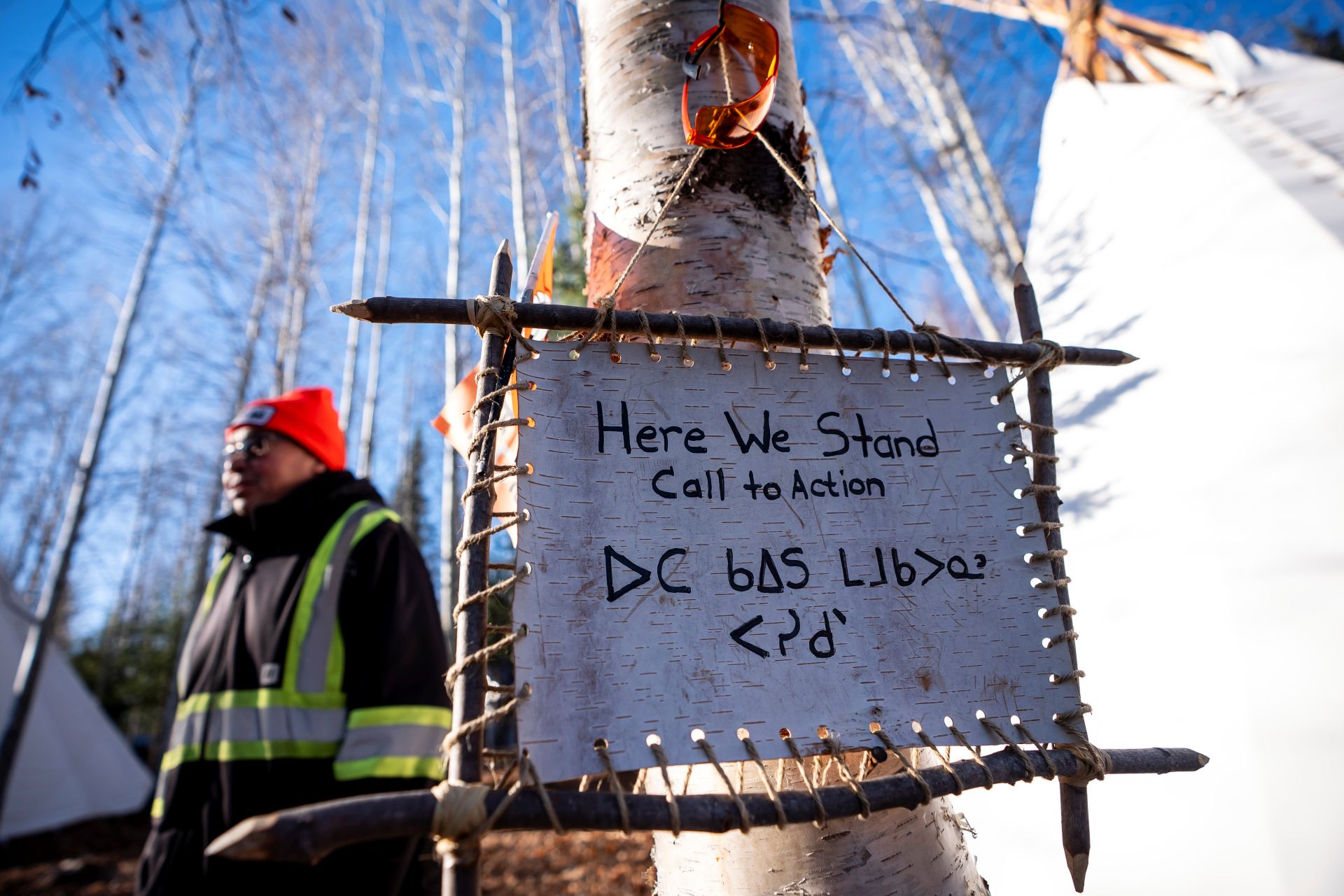 A man stands with a sign posted on a birch tree near his teepee. The sign says “Here we stand. Call to action” alongside some Indigenous script.