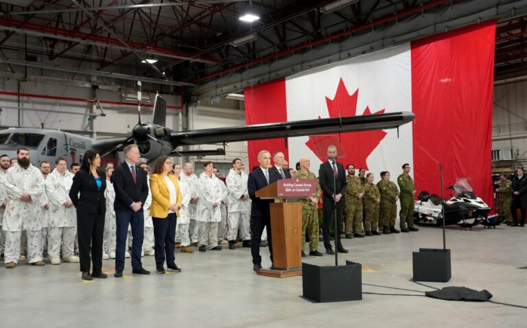 In a hanger, Carney stands in front of a long row of people in military fatigues and a grey aircraft.