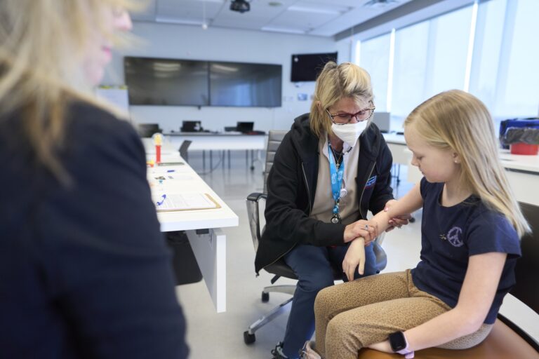 A masked nurse administers a shot to a blond girl’s right arm.