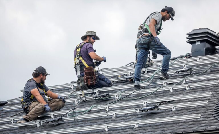 Three men sit, kneel or stand on the roof of a home covered in metal brackets.
