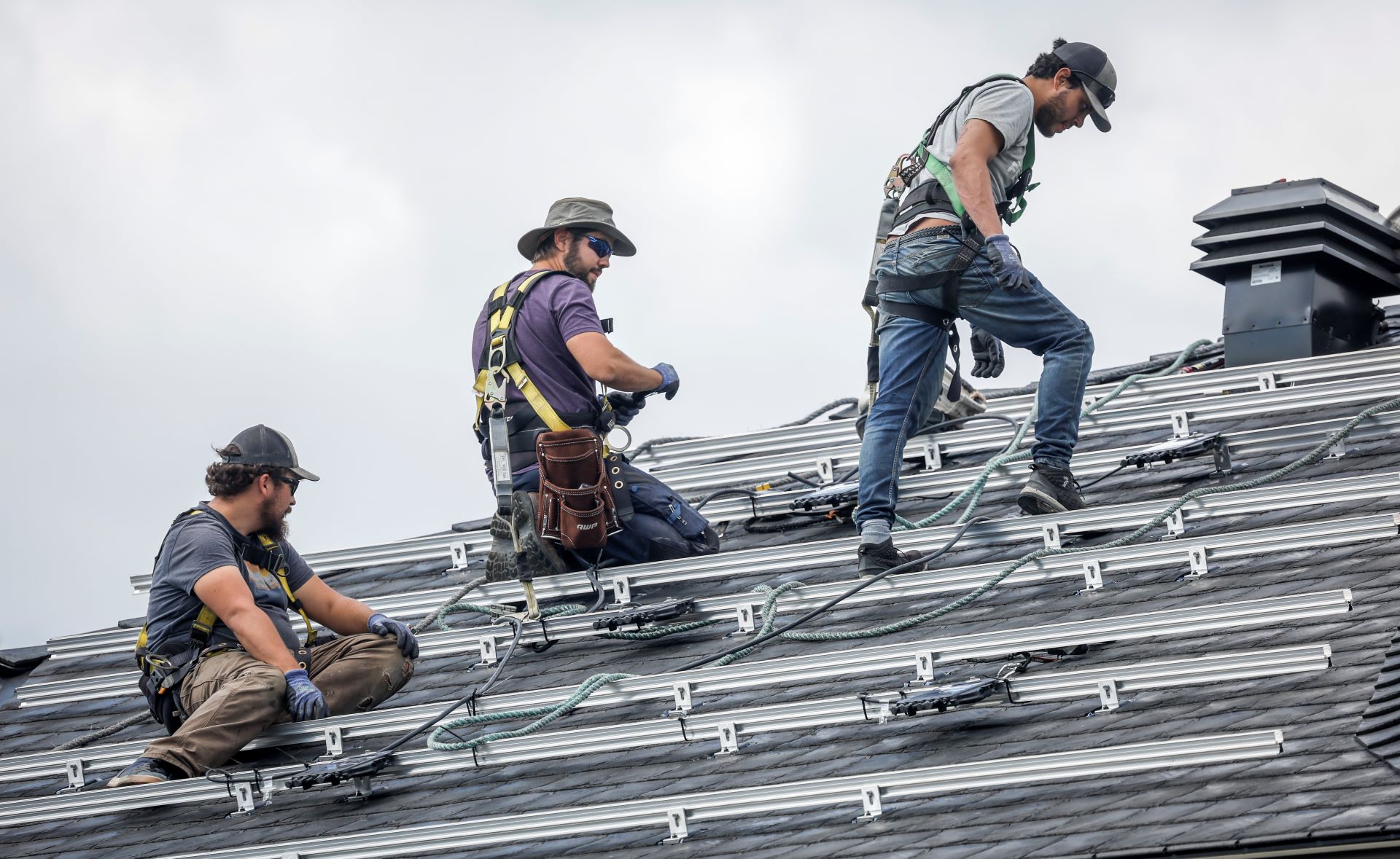Three men sit, kneel or stand on the roof of a home covered in metal brackets.