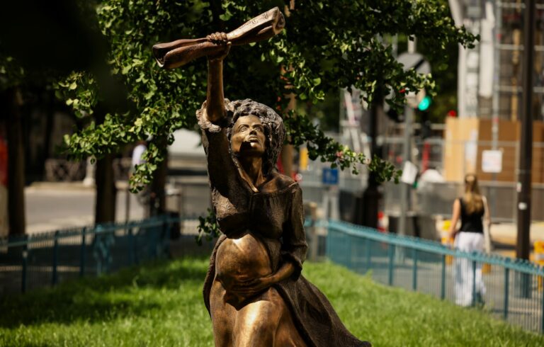Statue en bronze représentant une femme enceinte levant un fusil au-dessus de sa tête, installée dans un espace vert en milieu urbain, avec des arbres et des immeubles en arrière-plan.