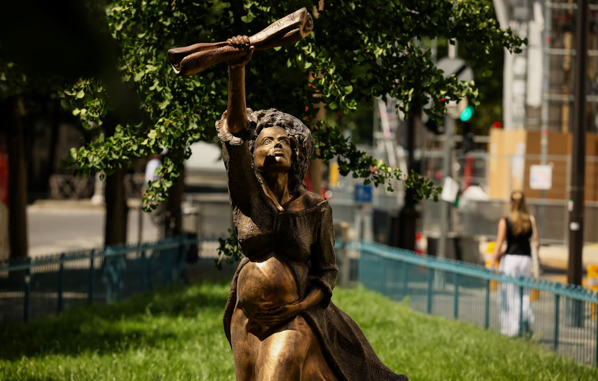 Statue en bronze représentant une femme enceinte levant un fusil au-dessus de sa tête, installée dans un espace vert en milieu urbain, avec des arbres et des immeubles en arrière-plan.