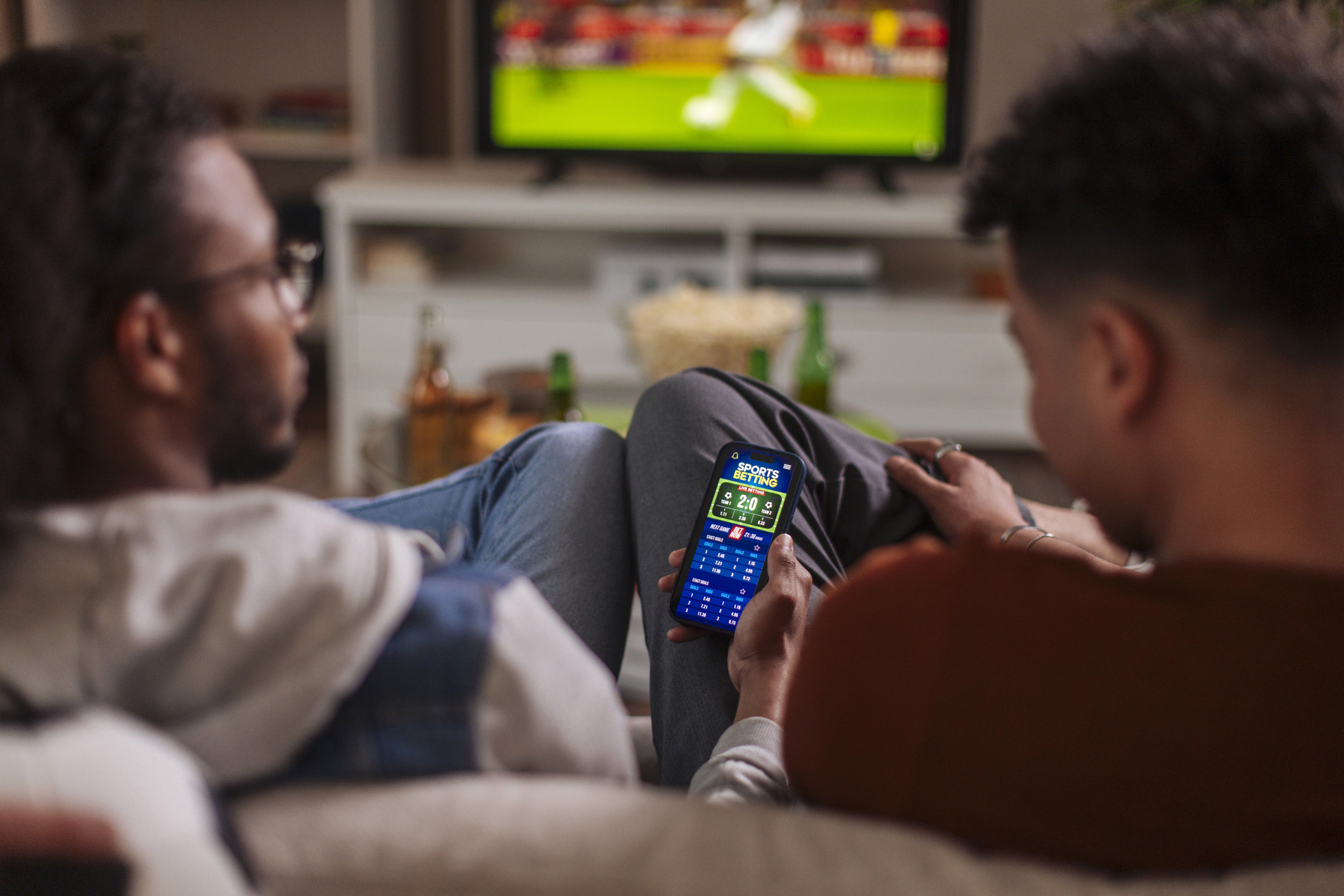 Two men look at a betting app on a telephone while watching soccer.