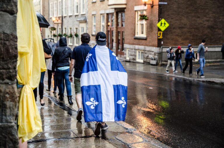 Une personne marche sur un trottoir en portant le drapeau du Québec sur son dos comme une cape.