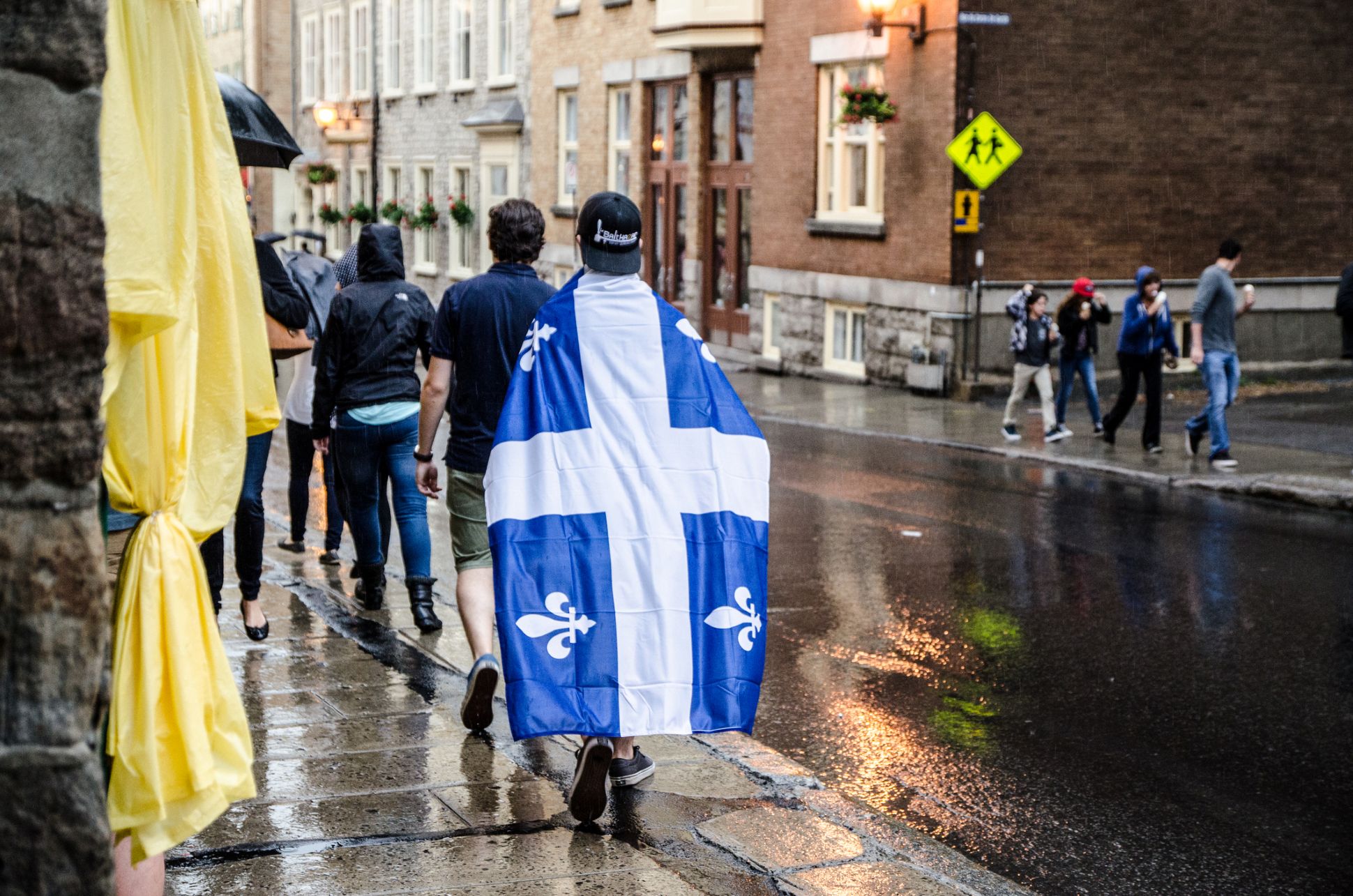 Une personne marche sur un trottoir en portant le drapeau du Québec sur son dos comme une cape.
