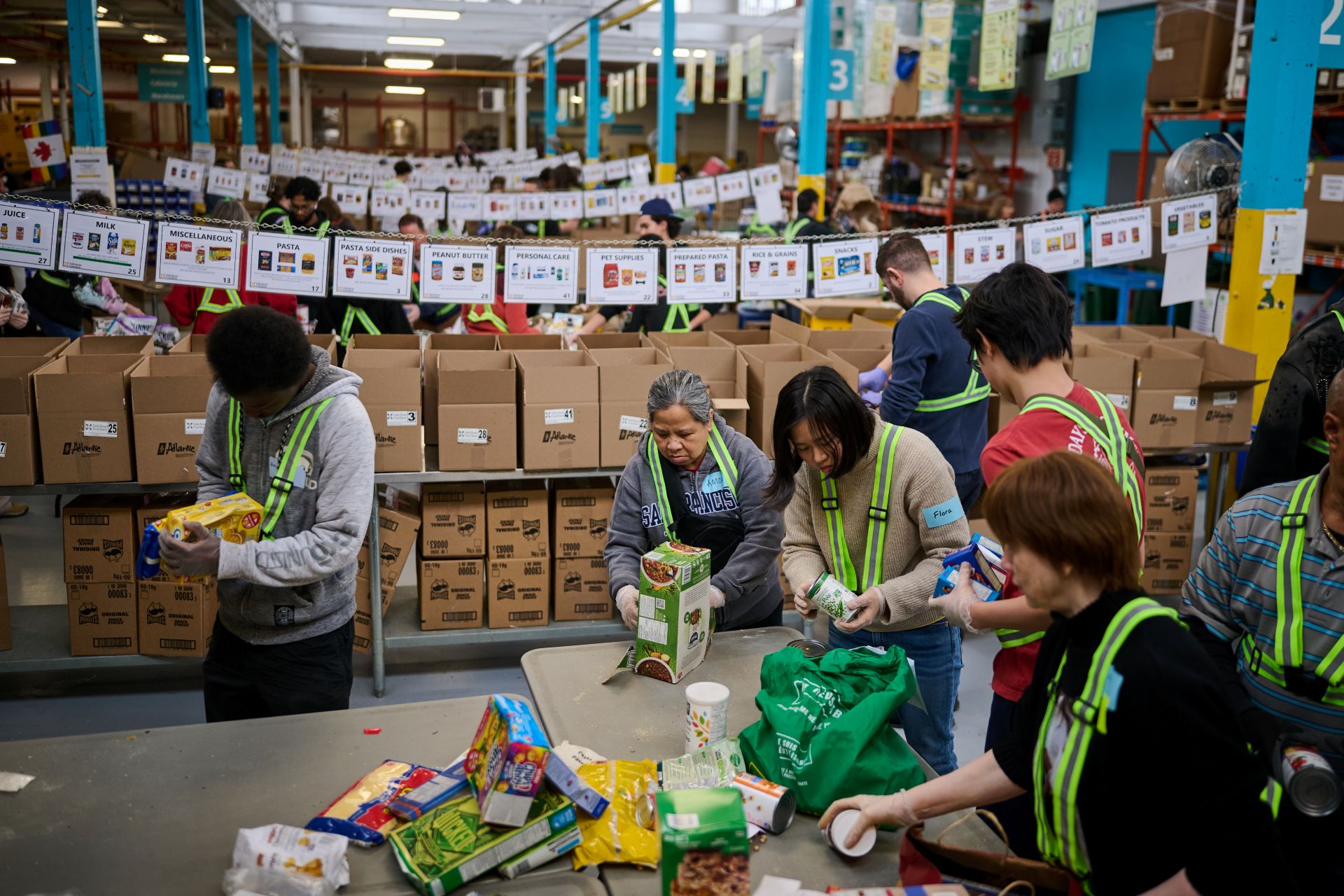 Volunteers wearing green safety vests sort and pack food donations into cardboard boxes at a food bank warehouse.