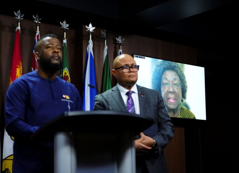 Two male black leaders stand at a podium while Jean Augustine appears on screen in the background.