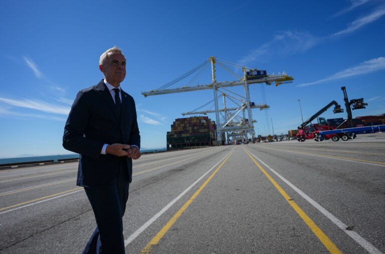 Carney walks across the loading area in front of gantry cranes and a container ship alongside the port facilities.