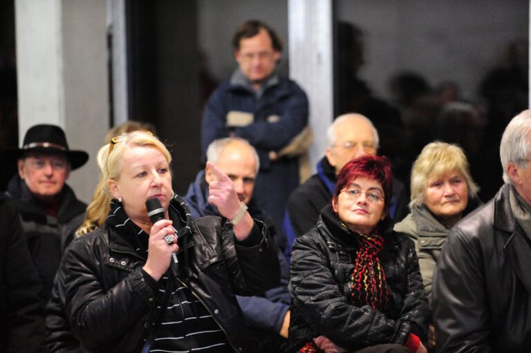A woman holding a microphone gestures with her hand as other citizens look on.