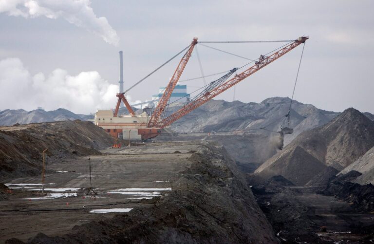 Une excavatrice à benne traînante dans une mine de charbon à ciel ouvert sous un ciel gris, avec une centrale thermique au charbon laissant échapper un panache de fumée en arrière-plan.