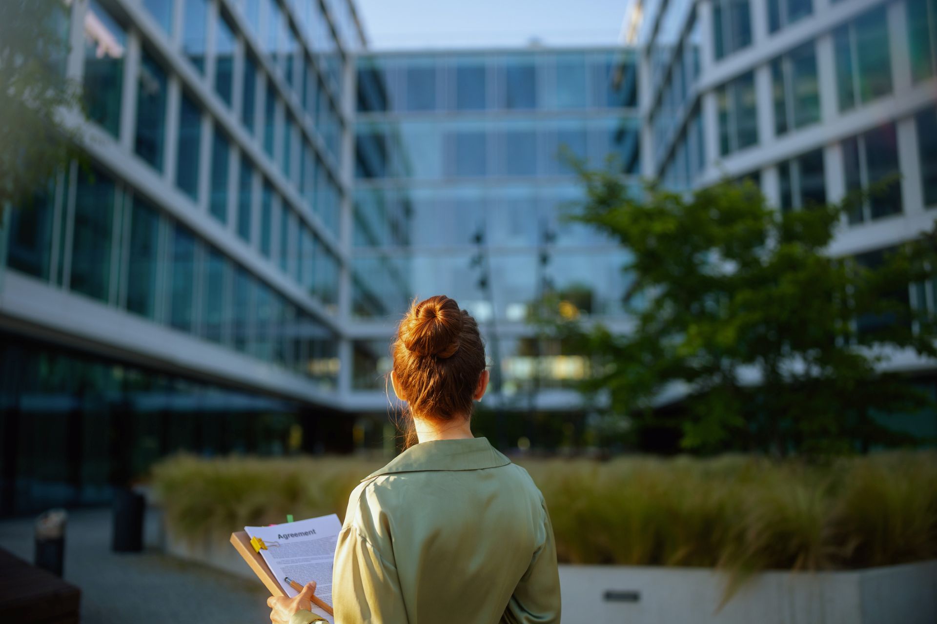 Une femme vue de dos avec à la main un document observe un espace vert entouré de bâtiments en verre.