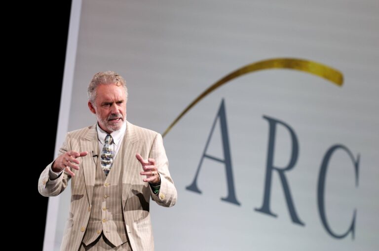 Jordan Peterson se tient sur scène devant le logo de l'Alliance pour une citoyenneté responsable. Il énonce un discours en gesticulant.