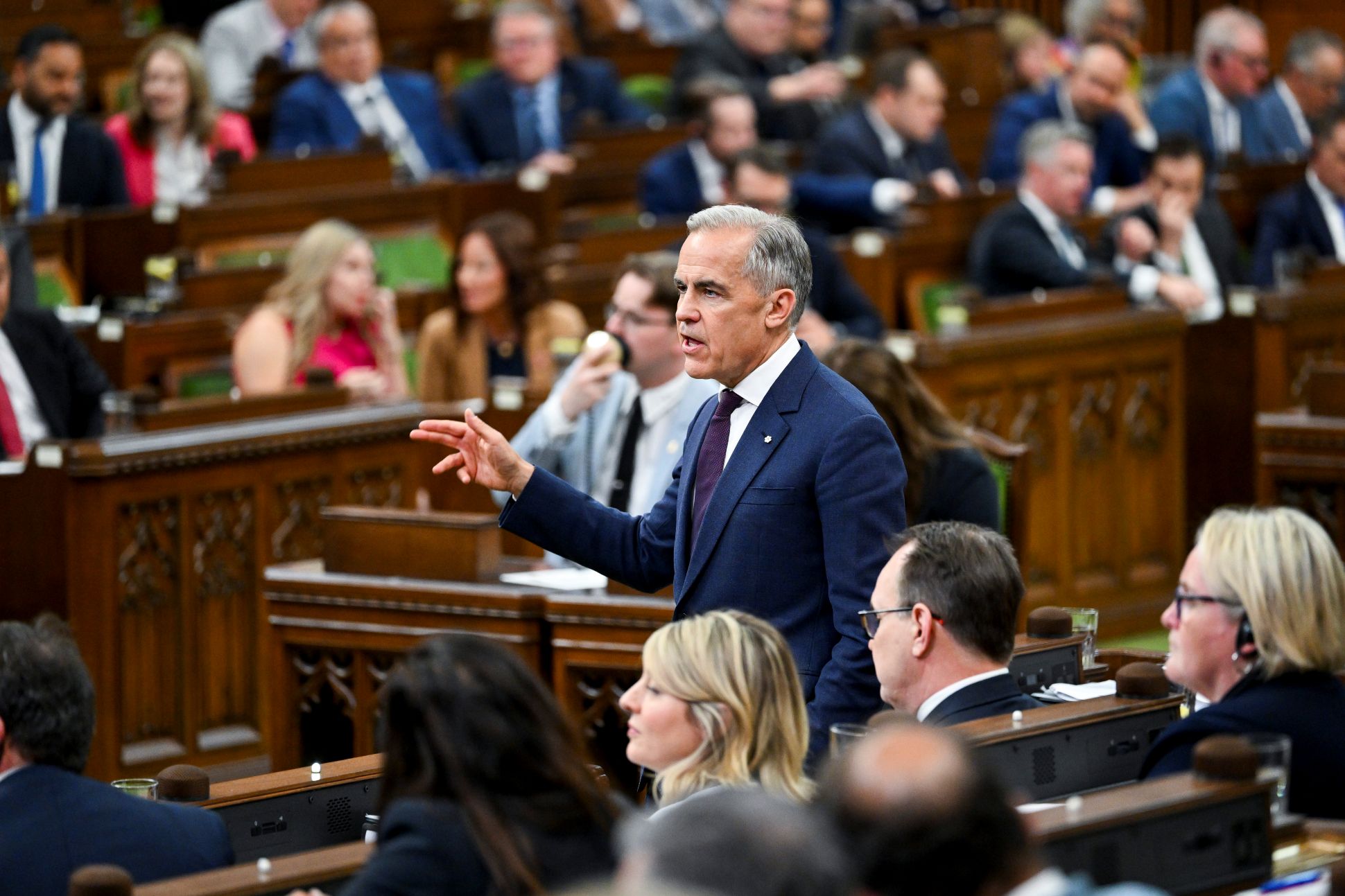 Mark Carney, wearing a blue suit, speaks in the House of Commons, surrounded by members of Parliament seated on the House benches.