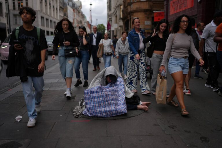 People rush past a man sitting on the street wrapped in a blanket.
