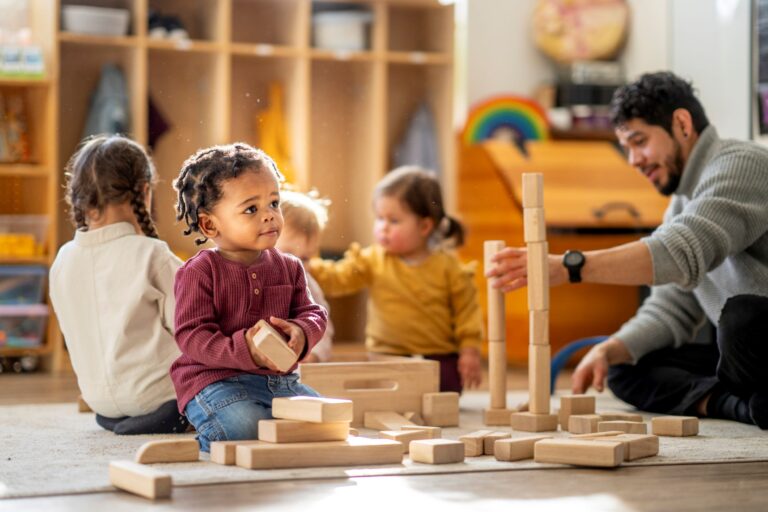 Un éducateur joue avec trois jeunes enfants qui construisent une structure avec des blocs de bois dans une salle de jeux d’un centre de la petite enfance.