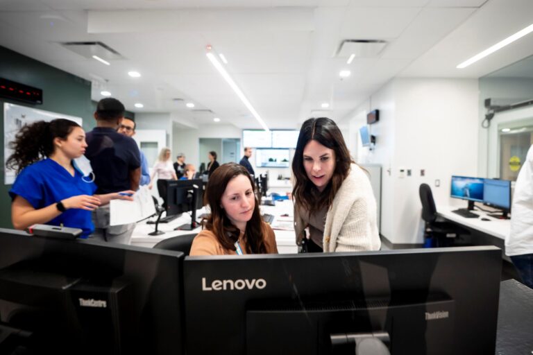 In an area filled with computer stations, two women look at a screen together.