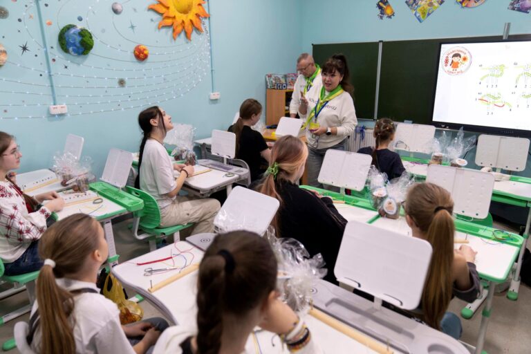 A dozen or so teen girls sit at desks in rows, listening to a teacher speaking.