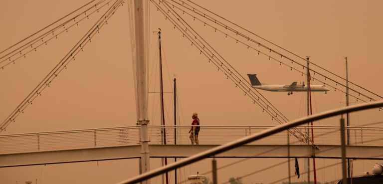 A bridge is in the foreground and a small passenger plane in the background amid an orange haze.