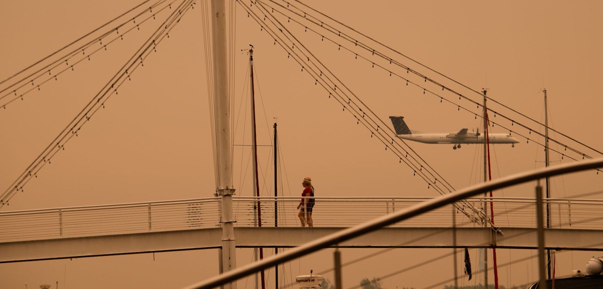 A bridge is in the foreground and a small passenger plane in the background amid an orange haze.