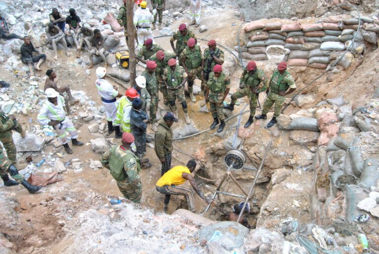A dozen men in fatigues stand around a small opening in the ground where a man is either coming out or going down.