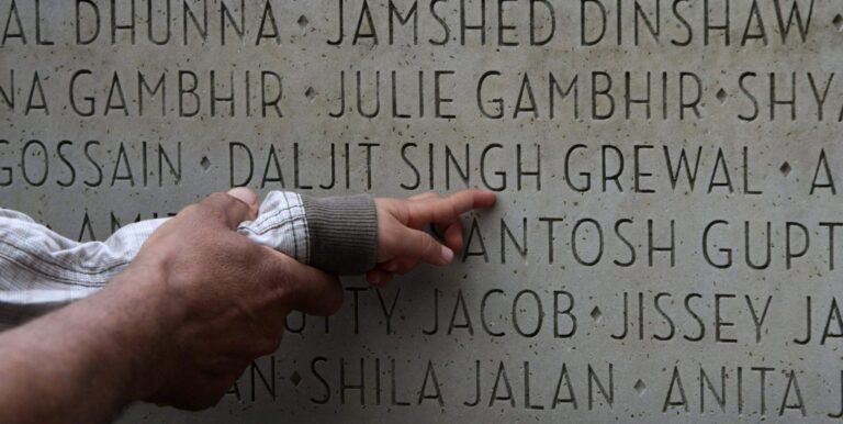 An adult’s hand holds a child’s wrist as the child points toward the name Daljit Singh Grewal on a stone memorial with hundreds of names.